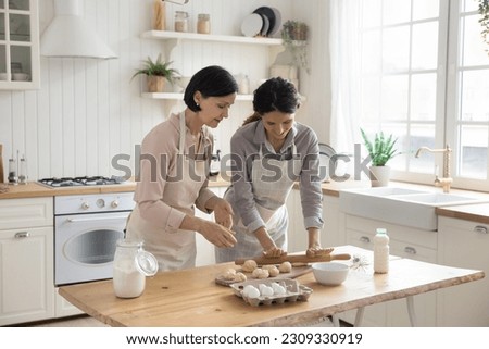 Positive adult daughter helping mature mom to bake, rolling raw dough on kitchen table, preparing homemade dessert, bakery meal, sharing domestic household chores