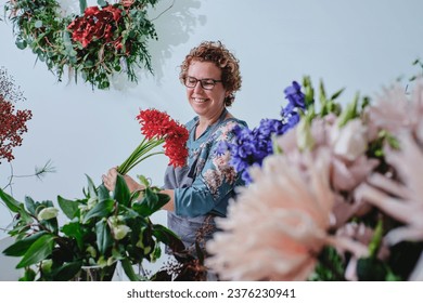 Positive adult female florist with short blond curly hair and glasses standing with bunch of red blooming flowers surrounded by various plants in floral shop - Powered by Shutterstock