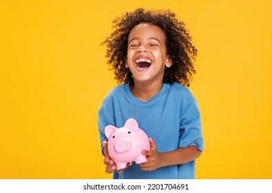 Positive African American Boy In Casual Outfit Laughing And Saving Money In Piggy Bank Against Yellow Background