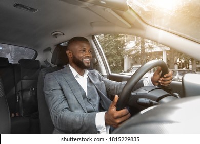 Positive African American Businessman In Expensive Suit Driving Car. Cheerful Young Black Handsome Entrepreneur Going To Business Meeting By His Luxury Car, Front Panel Shot, Copy Space, Sun Flare