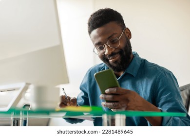 Positive african american businessman using cellphone and taking notes, making list or managing plans, sitting at workplace in office. Time management, business affairs concept