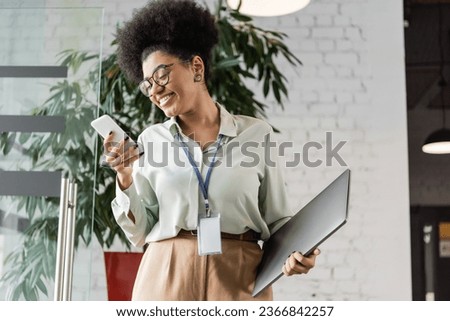 positive african american businesswoman in glasses holding laptop and using smartphone in office