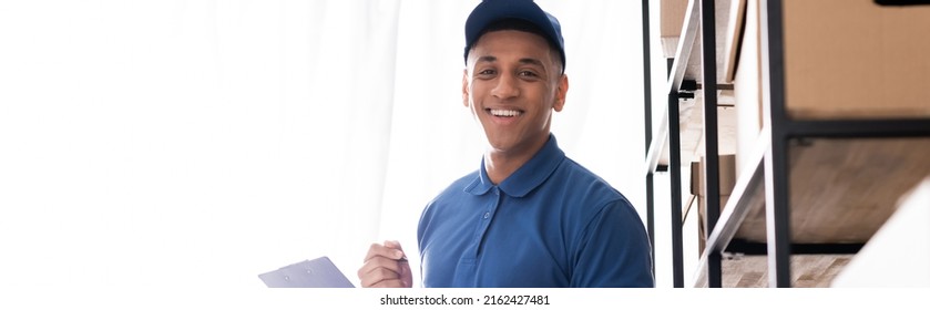 Positive African American Deliveryman In Uniform Holding Clipboard And Looking At Camera Near Boxes On Rack In Online Web Store, Banner