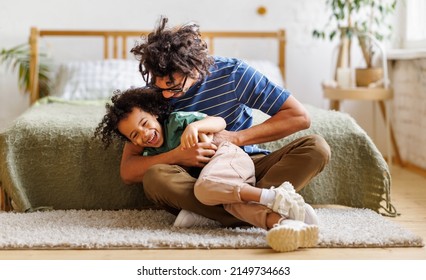Positive African American father tickling laughing son while sitting on floor in bedroom and having fun
