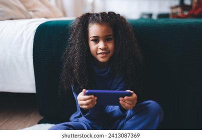 Positive African American female child with curly hair smiling and looking at camera while sitting on floor mat and using smartphone while watching program - Powered by Shutterstock