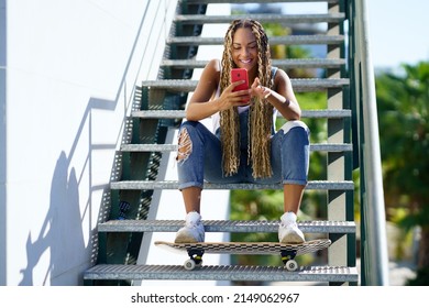 Positive African American Female Skater With Braids And Skateboard Sitting On Stairs And Messaging On Social Media On Cellphone In Summer