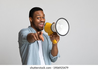 Positive african american guy shouting in megaphone and pointing at camera over grey background, copy space. Happy black man screaming with loudspeaker, cheering up, making advertisement