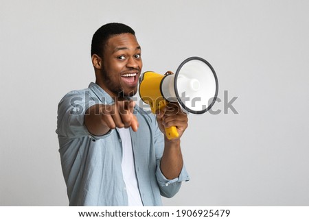 Positive african american guy shouting in megaphone and pointing at camera over grey background, copy space. Happy black man screaming with loudspeaker, cheering up, making advertisement