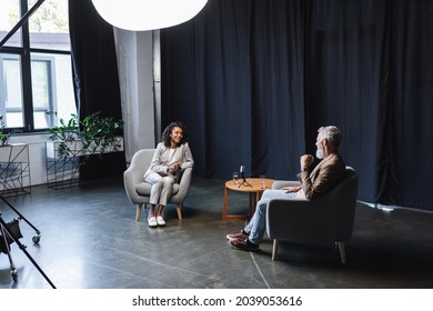 positive african american journalist in suit talking with businessman sitting in armchair during interview
