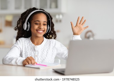 Positive African American Kid Cheerfully Waving At Laptop Screen, Using Wireless Headphones. Smiling Black School Girl Having Video Chat With Tutor, Enjoying Studying From Home, Kitchen Interior