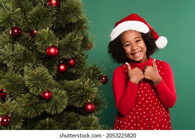 Positive african american kid in red santa hat showing thumbs up near christmas tree on green background - Powered by Shutterstock