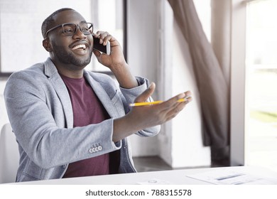 Positive African American Male Enterpreneur Dressed In Formal Clothes, Has Conversation Over Smart Phone, Gestures Actively With Hand Holds Pencil, Indicates At Something As Speaks With Friend