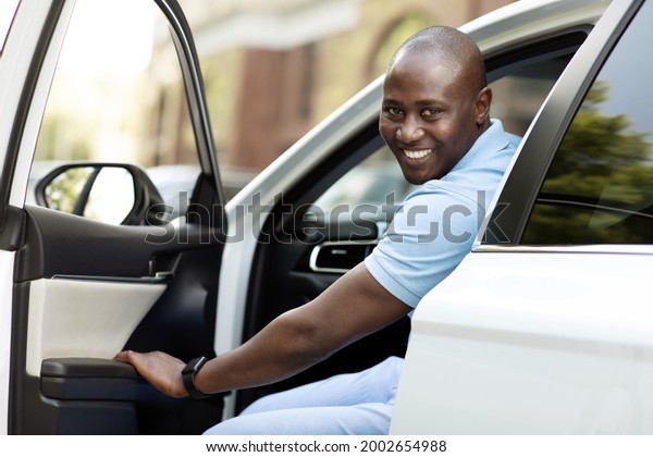 Positive african american man getting in his\
brand new car, closing automobile door and smiling at camera. Happy\
black guy in casual outfit going summer vacation by auto, car trip\
concept
