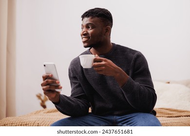 Positive african american man holding coffee and smartphone in bedroom - Powered by Shutterstock