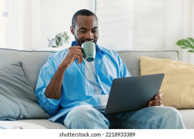 Positive african american man sitting on couch, drinking coffee and working on laptop computer, copy space. Black guy checking email. Handsome male freelancer using computer while drinking hot tea - Powered by Shutterstock