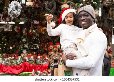 Positive African American Man With Small Daughter In Hats Buying Decoration At Christmas Fair Outdoor