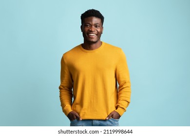 Positive african american millennial guy in yellow sweater and jeans holding hands in pockets and cheerfully smiling at camera over blue studio background, copy space. Millennials lifestyle - Powered by Shutterstock