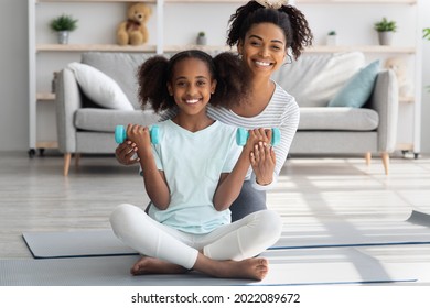 Positive African American Mother And Daughter With Barbells Sitting On Fitness Mat In Living Room, Athletic Black Woman Showing Her Teen Kid Working Out With Dumbbells, Home Interior, Copy Space