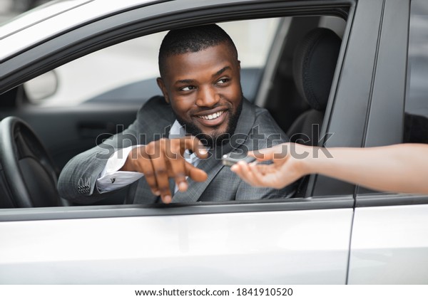 Positive african\
american young businessman sitting in brand new car, taking\
automatic key from car dealer hands. Smiling black man in suit\
taking his car from parking,\
closeup