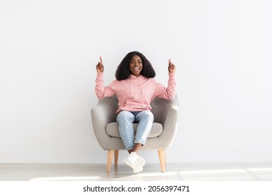 Positive african american young lady sitting in arm chair over white wall at home, pointing up at copy space for text or advertisement, happy black woman recommending something exciting