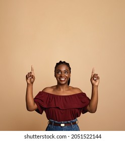 Positive african american young woman pointing with both hands and looking up at copy space, happy millennial black lady showing empty space for advertising over beige studio background