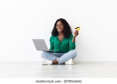 Positive African American Young Woman Sitting On Floor Next To White Wall, Holding Credit Card And Notebook, Banking Online, Happy Black Lady Shopping On Internet, Using Laptop, Copy Space