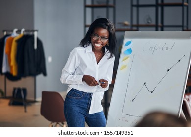 Positive African Business Lady In White Shirt And Blue Jeans Stand Talking Explaining Making Flip Chart Presentation For Younger Leaders, Share Business Experience With Smile