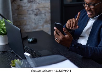 Positive African businessman using smartphone presses his finger on screen of a mobile phone, browsing the Internet or checking an Internet application on a smartphone while sitting at an office desk