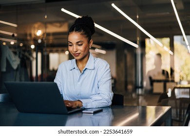 Positive African businesswoman sitting by the desk, typing something on the laptop.