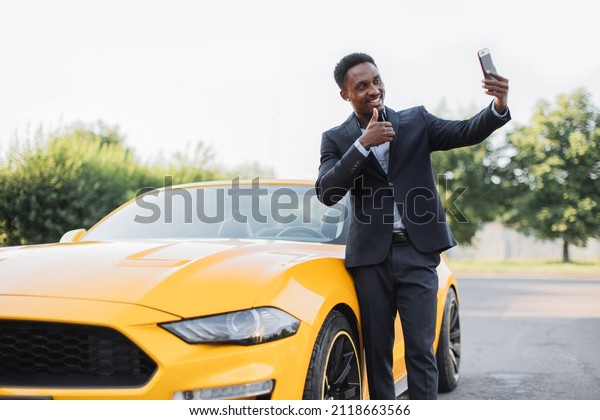 Positive african man in suit having video\
call on smartphone while standing near luxury sport car outdoors.\
Businessman standing near yellow auto, smiling and waving during\
online conversation