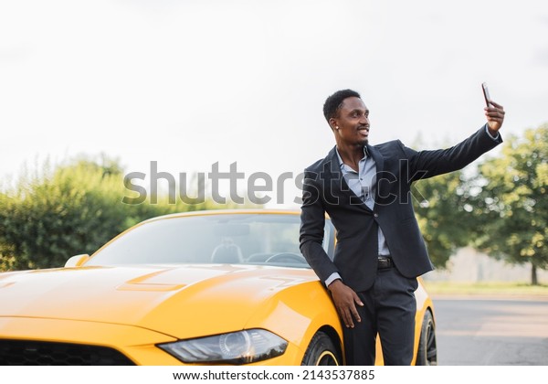 Positive african man in suit having video\
call on smartphone while standing near luxury sport car outdoors.\
Businessman standing near yellow auto, smiling and waving during\
online conversation