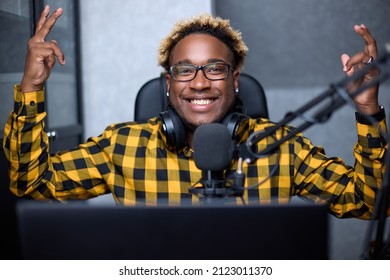 Positive African Man Wearing Headphones Records Or Broadcasts A Podcast Using A Microphone In His Small Broadcast Studio. A Black Man In Glasses And A Plaid Shirt Threw Up His Hands Emotionally