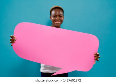 Positive african woman with short hair holding pink speech bubble with copy space for text. Beautiful, cheerful, smiling girl with cardboard speech bubble posing on blue background in studio.