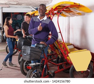 Positive African-American man pedicab driver standing near rickshaw cycle