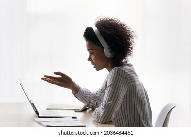 Positive afro american teen female wear earphones sit at desk by laptop learn on distance talk speak ask answer questions at virtual consultation. Black gen z lady confer online in app for video calls - Shutterstock ID 2036484461