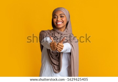 Positive afro muslim girl in hijab gesturing thumbs up and smiling at camera while posing over yellow background in studio, empty space