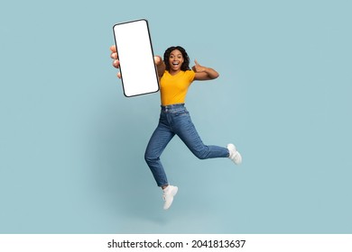 Positive afro-american woman jumping up and demonstrating big smartphone with blank screen, smiling and showing thumb up, showing newest mobile application, mockup, blue studio background, copy space