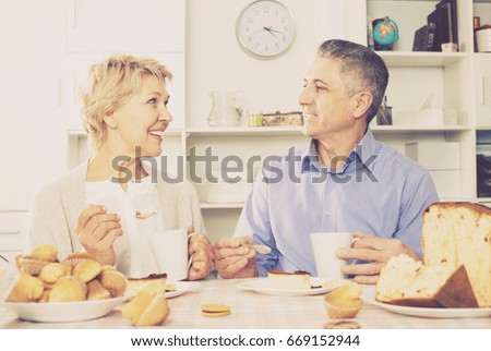 Positive aged married couple have breakfast and have coffee with cakes at table in house