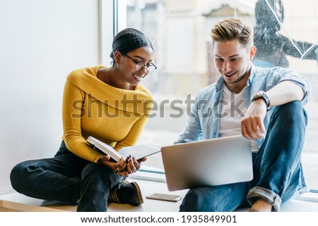 Positive ambitious multiracial undergraduates using laptop together while reading article enthusiastically during exam preparation sitting against window in university library