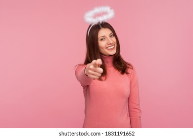 Positive angelic brunette woman in pink sweater with nimbus smiling pointing finger to camera, choosing lucky winner. Indoor studio shot isolated on pink background