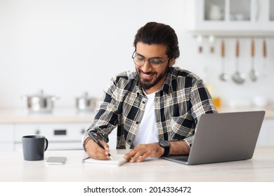 Positive Arab Guy With Glasses Looking For Job Online, Sitting At Table, Using Laptop And Taking Notes, Kitchen Interior, Copy Space. Excited Indian Man Student Working Online From Home, Remote Job