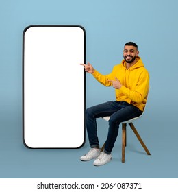 Positive Arab Guy Pointing At Big Smartphone With White Screen While Sitting On Chair Over Blue Background. Man Demonstrating Free Space For Your App Or Website Design, Mockup