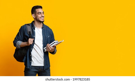 Positive Arab Guy Student With Backpack And Bunch Of Books Looking At Copy Space For Advertisement Over Yellow Studio Background, Panorama. Education, University, College, Studying, Course Concept