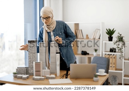 Positive arabian lady in hijab and glasses arranging architectural models of city complex placed on office desk near laptop and tablet. Talented professional developing urban area project at work.