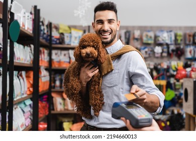 Positive arabian man holding poodle and looking at camera while paying with credit card in pet shop  - Powered by Shutterstock