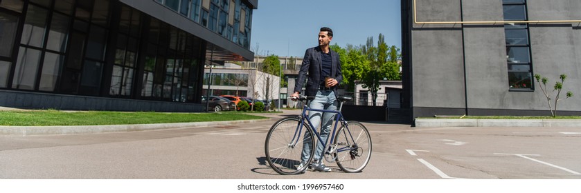 Positive arabian man with paper cup and bike standing near building on urban street, banner