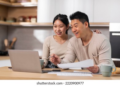 Positive asian couple handsome middle aged man and pretty young woman sitting at kicthen desk in front of laptop, holding bills, paying for goods and services on Internet from home, copy space - Powered by Shutterstock