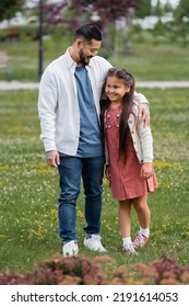 Positive Asian Dad Hugging Daughter On Meadow With Flowers In Park