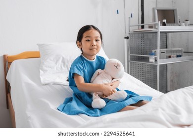 positive asian girl embracing toy bunny and looking at camera while sitting on hospital bed - Powered by Shutterstock