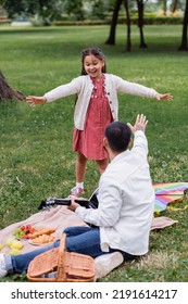 Positive Asian Girl Looking At Dad Holding Acoustic Guitar Near Food On Blanket In Park